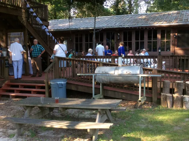 People gathered on a wooden porch with a picnic table and grill in a wooded outdoor setting