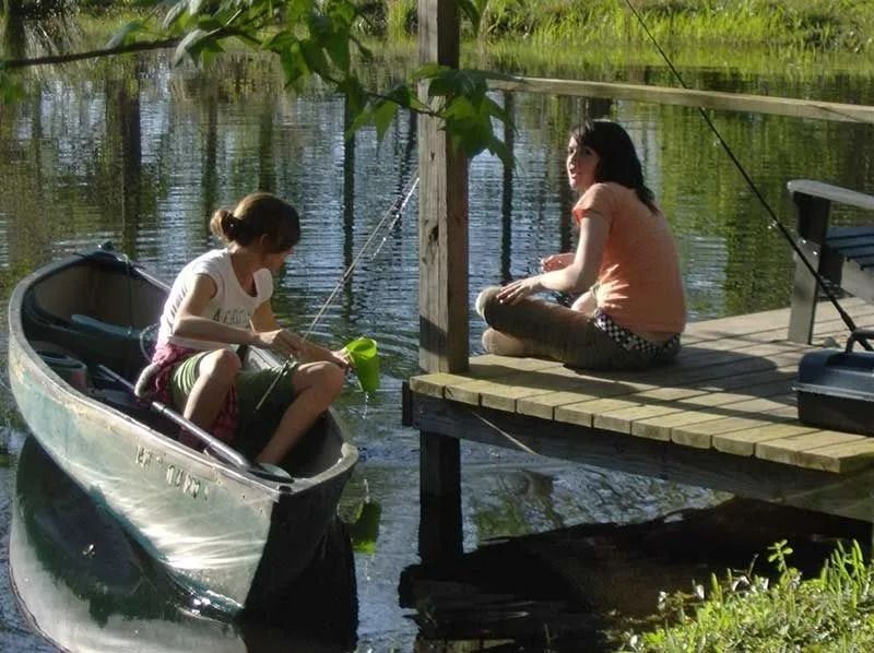 Two people sit by a canoe on a dock beside a calm pond in a sunny park.