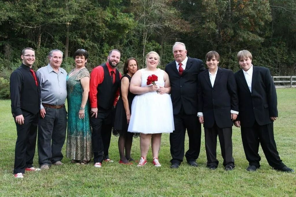 Group of people posing outdoors on grass, dressed in formal clothes for a celebration, with a bride in a white dress.