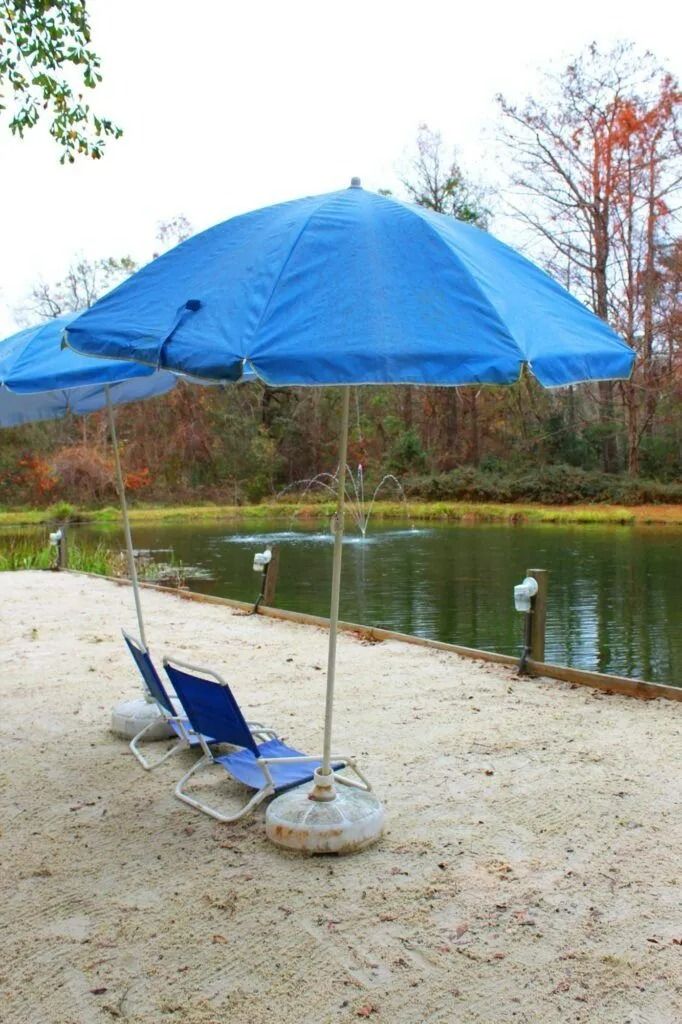 Blue beach umbrella and lounge chairs on a sandy lakeshore beside calm water, with trees in the background