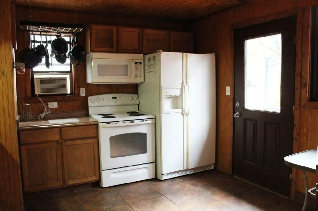 Small rustic kitchen with wood-paneled walls, white stove and fridge, microwave, and window air conditioner.