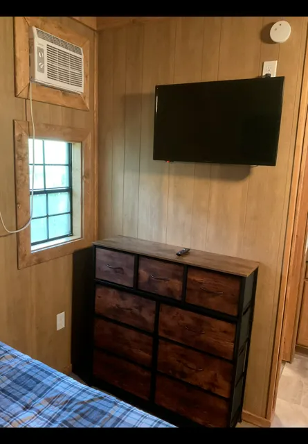 Small wood-paneled bedroom with wall-mounted TV, dresser, window, and a bed edge in the foreground