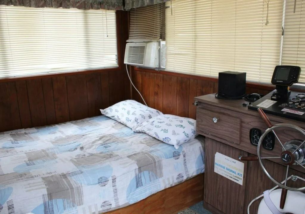 Small cabin bedroom with a single bed, wooden walls, and a desk with a fan and monitor.