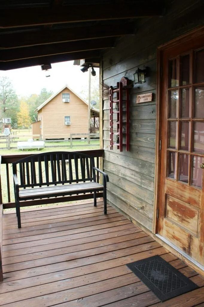 Covered wooden porch with bench, wood siding, glass door, and a cabin visible across the yard