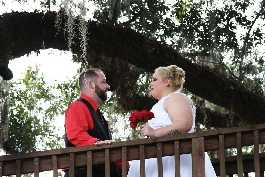 Couple facing each other on a wooden bridge, holding red flowers in a tree-lined outdoor setting