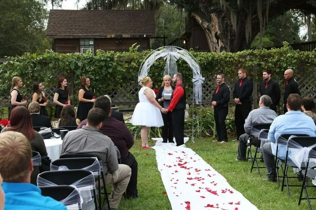 Outdoor wedding ceremony with bride and groom under a white arch, guests seated along a flower-strewn aisle.