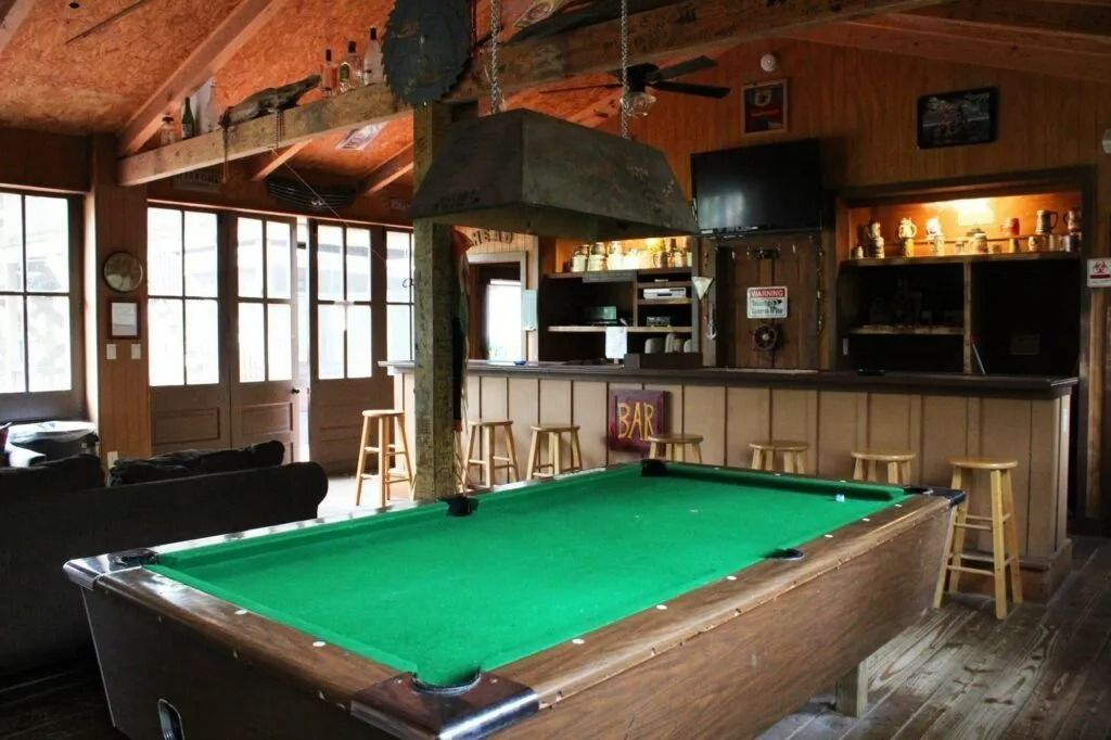 Rustic bar with a green pool table, wooden beams, stools, and a counter in the background