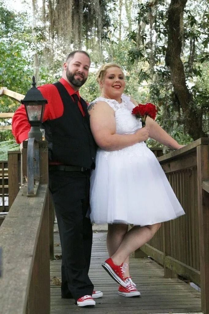 Couple posing on a wooden bridge; man in black and red, woman in white dress holding red flowers.