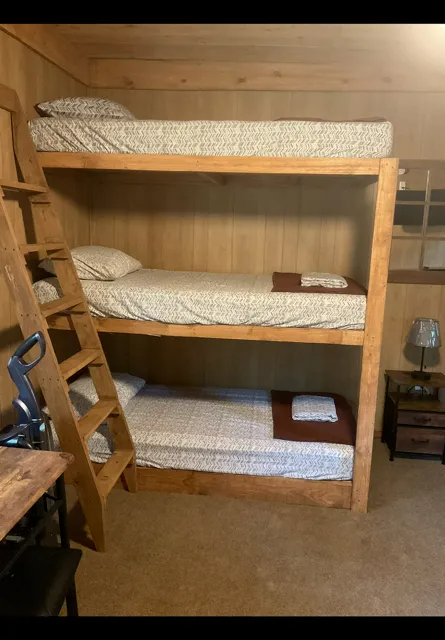 Wooden bunk bed with three beds in a rustic room, with a ladder on the left and a small table on the right