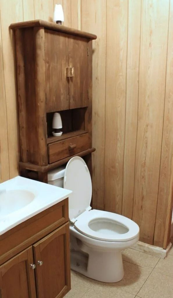 Small bathroom with wood-paneled walls, toilet, vanity, and a tall wooden cabinet above the toilet.
