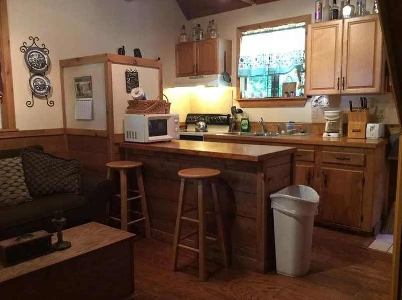 Small kitchen with wood cabinets, fridge, microwave, stools, sink, and a living area corner.