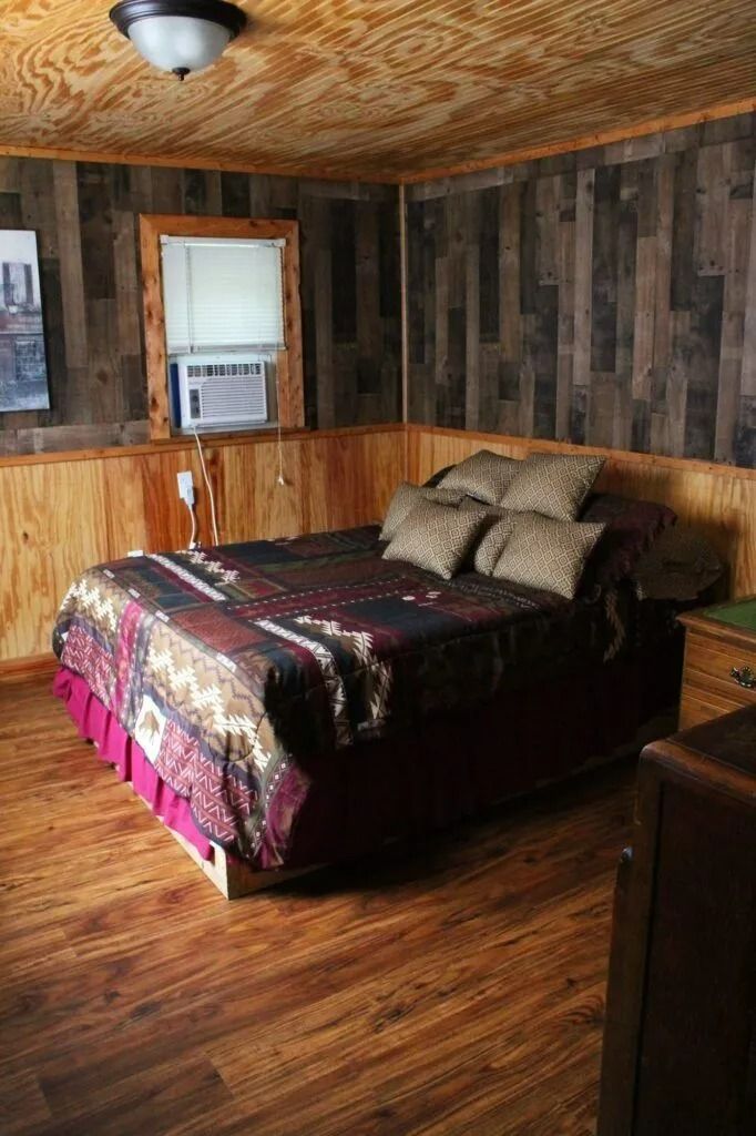 Small bedroom with wood-paneled walls, hardwood floor, a bed by the window, and patterned bedding.