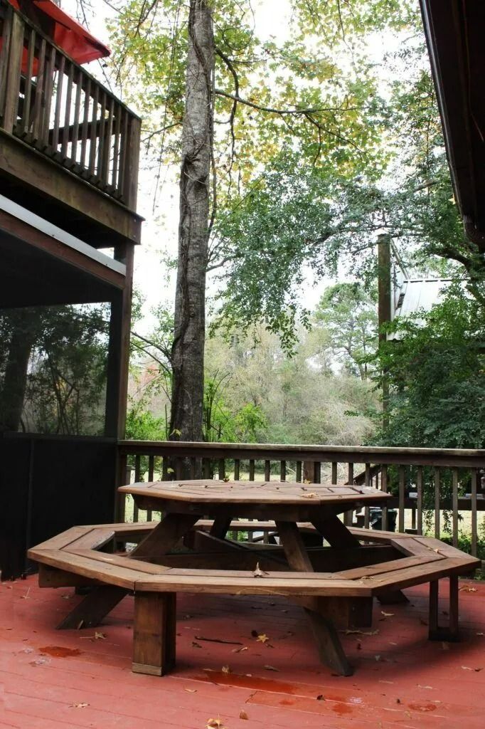 Wooden picnic table on a red deck beside a forest, with a balcony railing on the left.