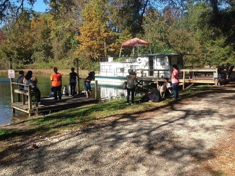 People standing by a dock and small white boat on a sunny riverbank with autumn trees.