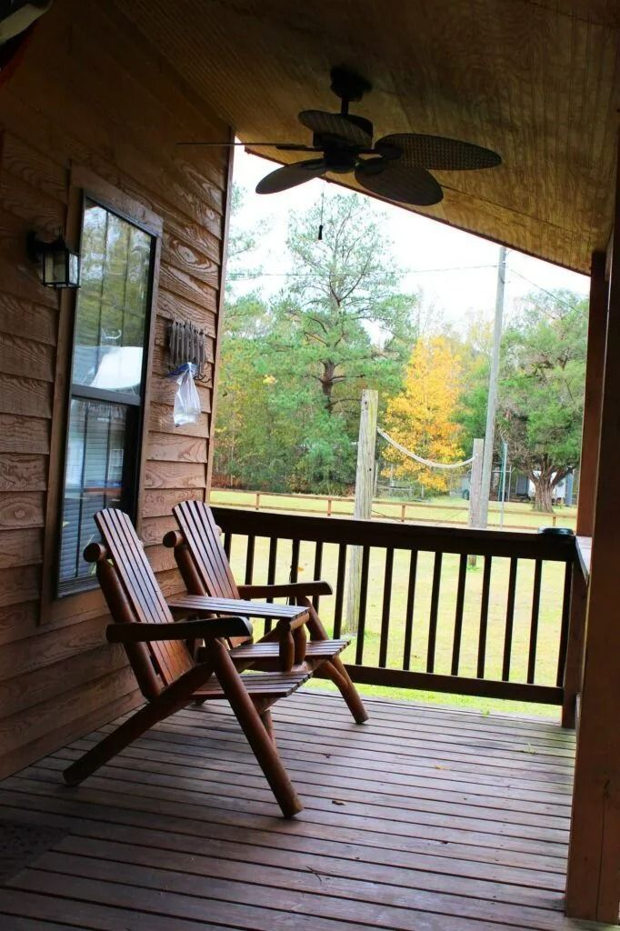 Wooden porch with two chairs, ceiling fan, and view of trees and yard through the railing.