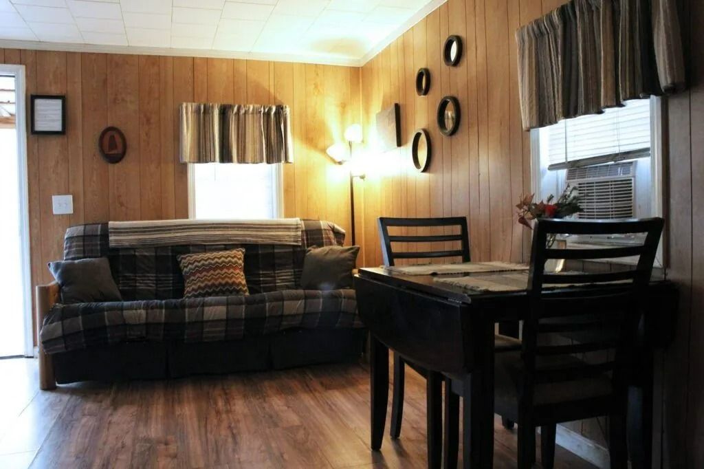 Cozy wood-paneled living room with sofa, dining table, wall decor, and windows with curtains