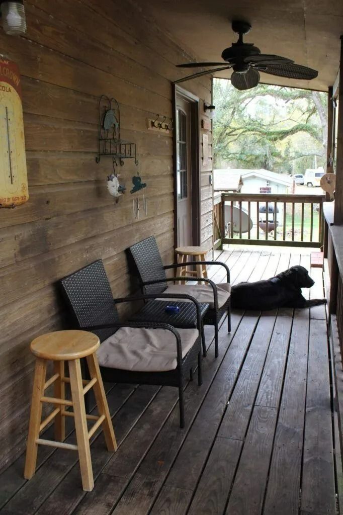 Covered porch with rocking chairs, stool, ceiling fan, and a black dog lying on the wooden floor.