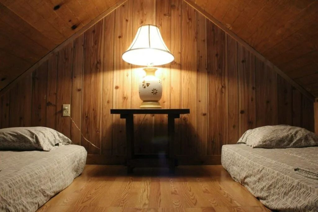 Cozy attic bedroom with twin beds, a bedside table, and a lamp centered on wood-paneled walls.