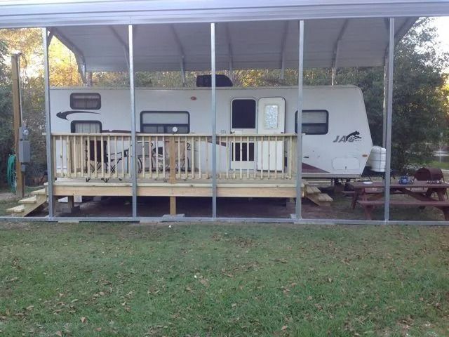 Camper trailer under a metal carport with a wooden porch in a grassy yard