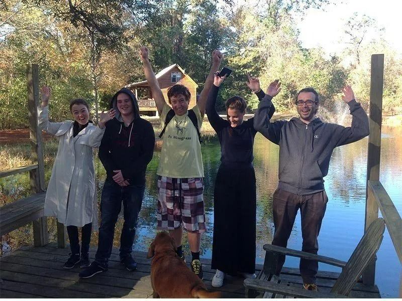 Five people standing outdoors with arms raised, smiling in a wooded area near a wooden deck.