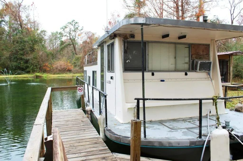 White houseboat docked on a wooden pier beside a calm lake with trees in the background