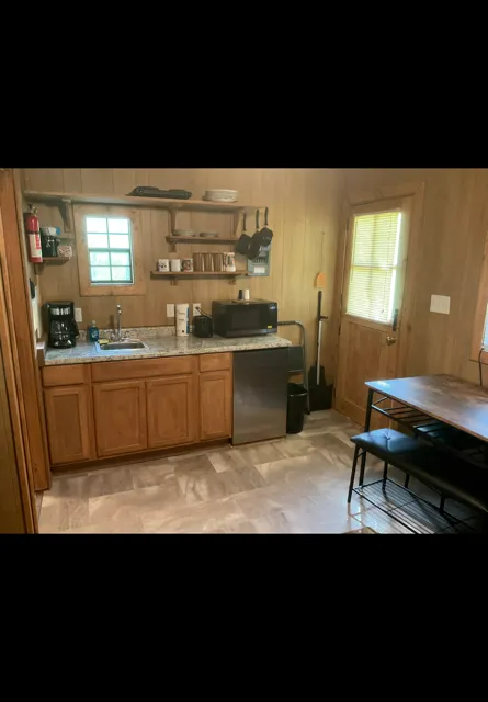Small rustic kitchen with wood cabinets, sink, stove, window, and a dining table by the door