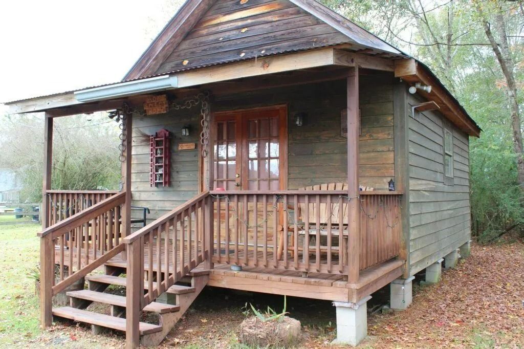 Small rustic wooden cabin with a porch, stairs, and railing in a wooded setting