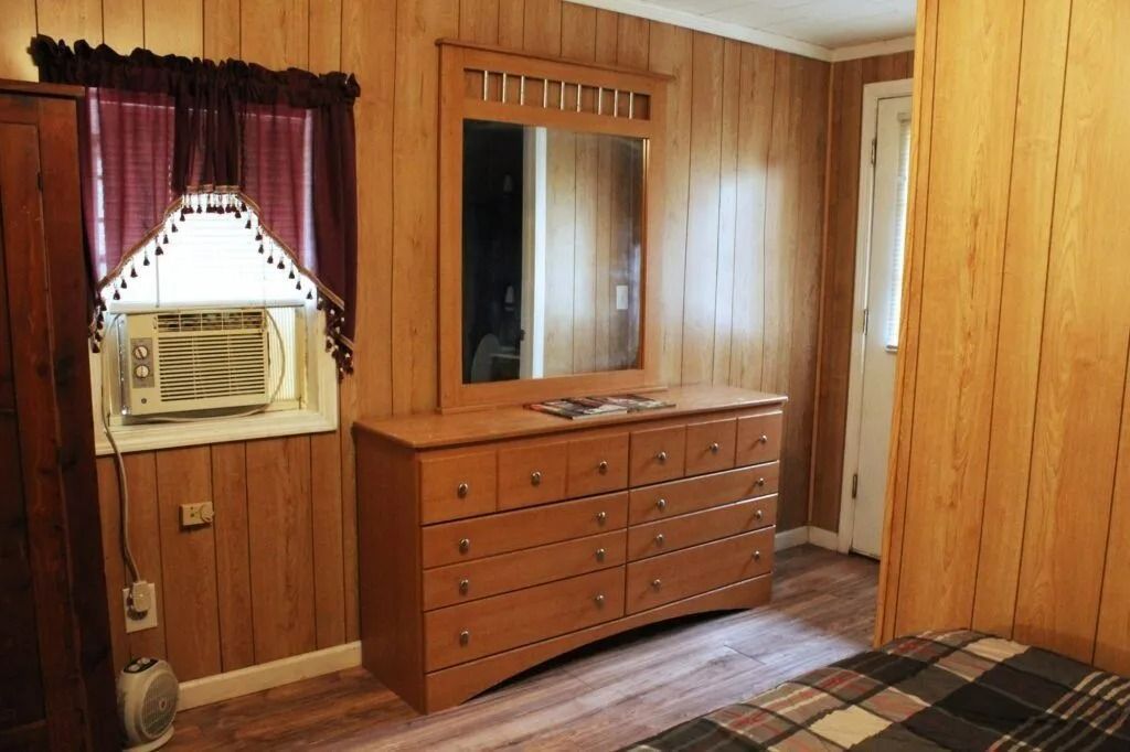 Wood-paneled bedroom with a dresser, mirror, window curtains, and plaid bedspread