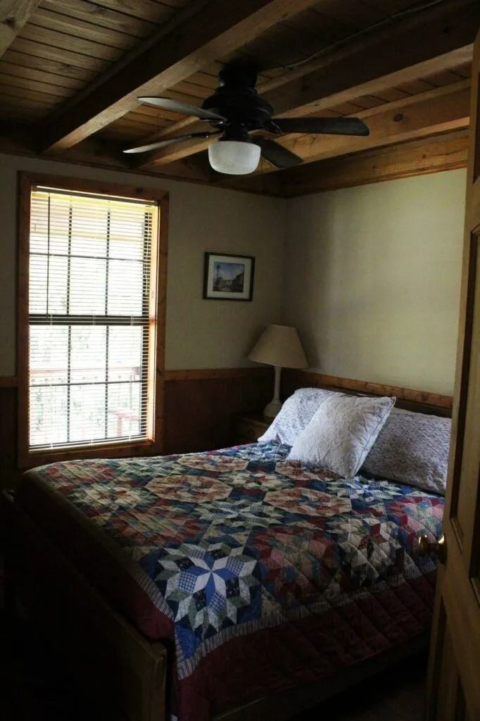 Small cozy bedroom with a quilted bed, ceiling fan, window blinds, and warm wood-paneled ceiling.