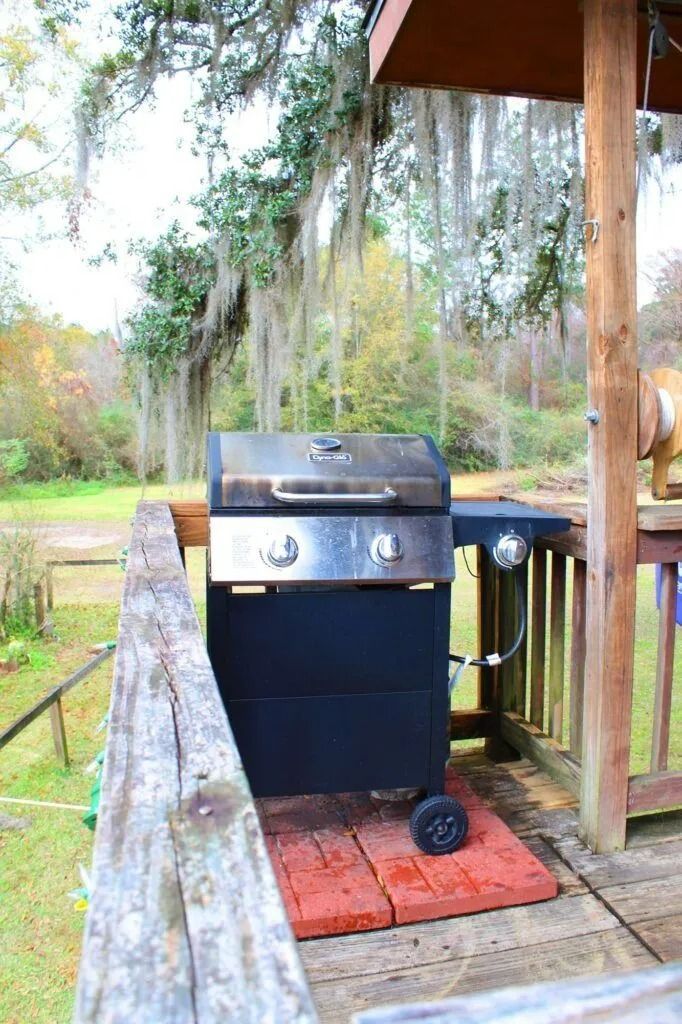 Black grill on a wooden deck beside a railing, under trees outdoors.