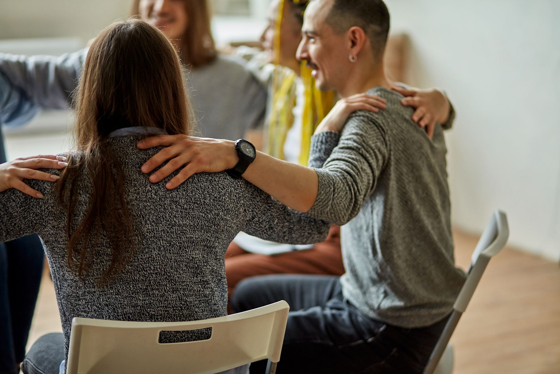 Group of people in a circle, arms around each other's shoulders; one person sits on a chair, smiling.