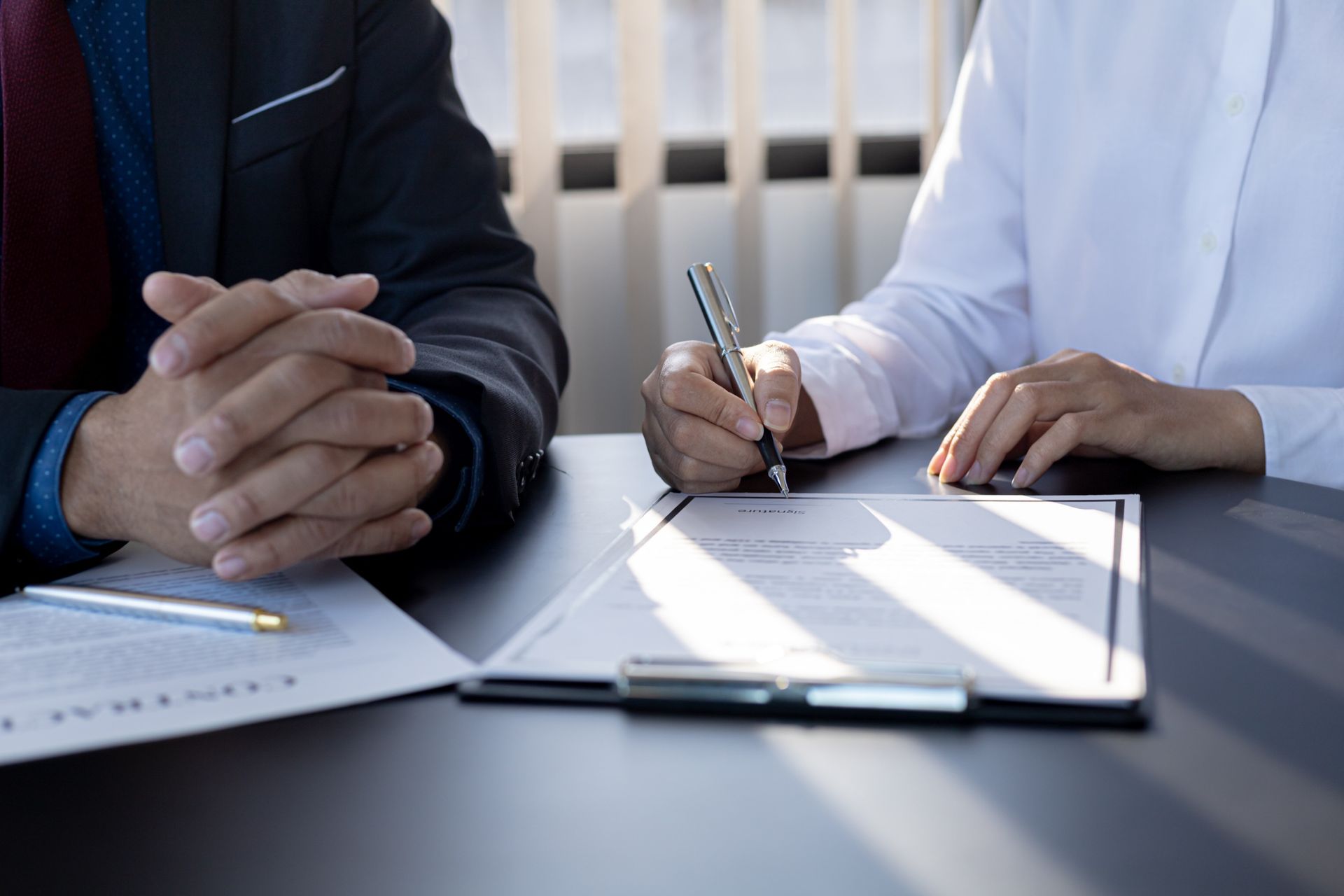 Person signing a document on a desk, next to another person with hands clasped.