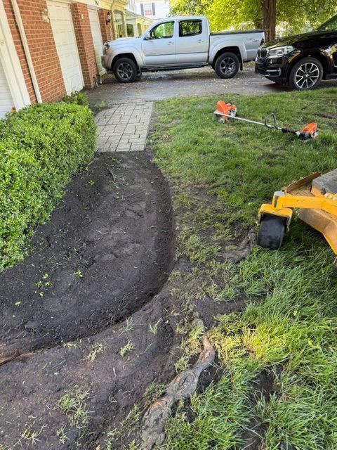 Freshly dug curved trench in a front yard beside a house, with a shovel and pickup trucks nearby