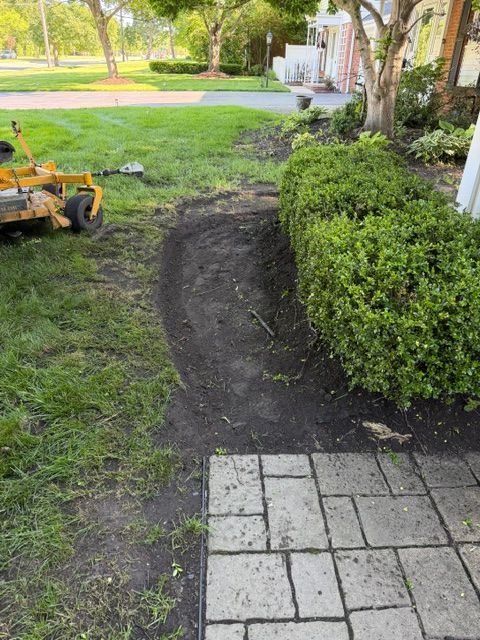 Garden path area with freshly dug soil beside a hedge and patio pavers, with a small yellow machine on the grass.
