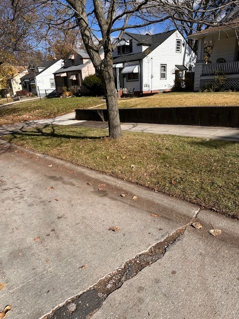 A residential street scene with houses, trees, grass, and a cracked sidewalk on a sunny day.