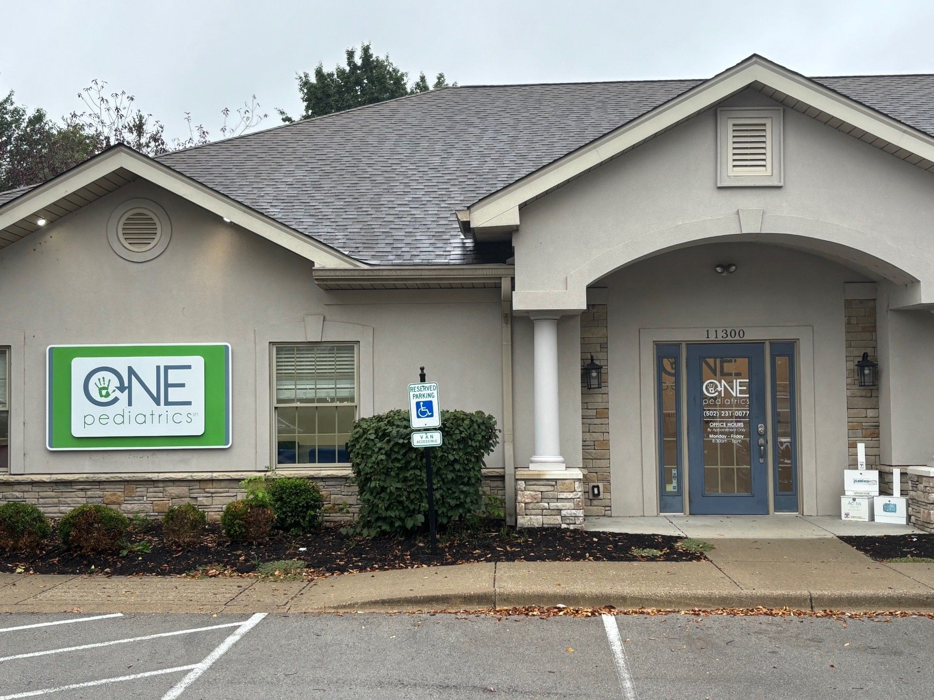 Exterior view of a light beige building with a green sign that reads "ONE pediatrics". The entrance has a blue door.