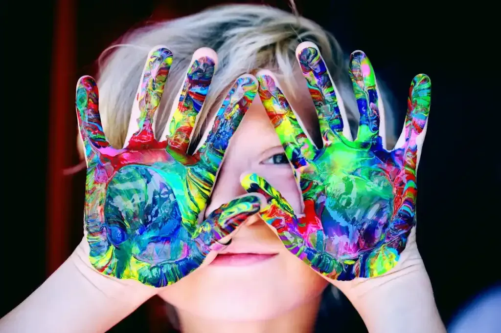 Child with hands covered in colorful paint, eyes peeking through fingers.
