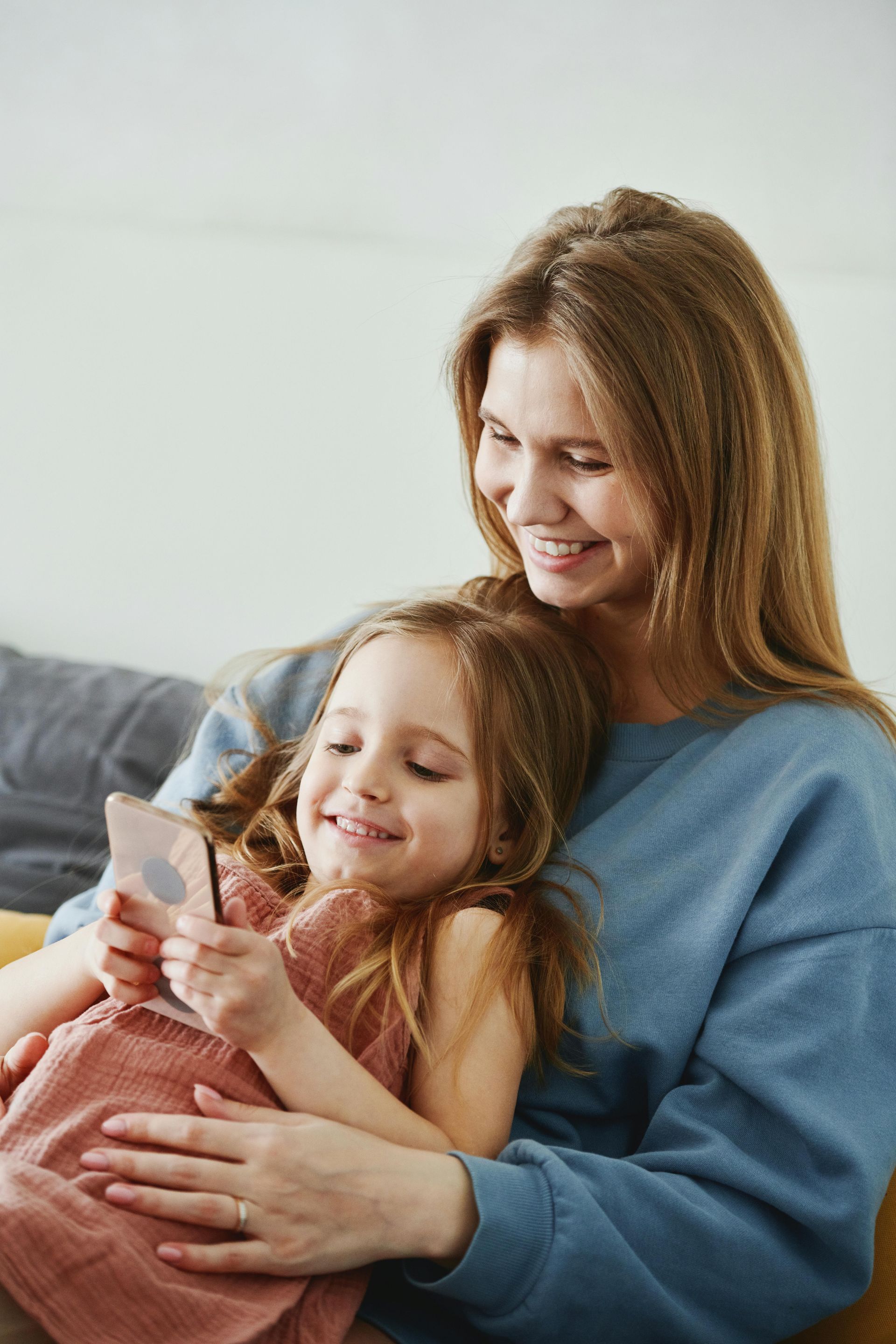 Woman and child smiling, looking at a phone held by the child, seated on a couch.