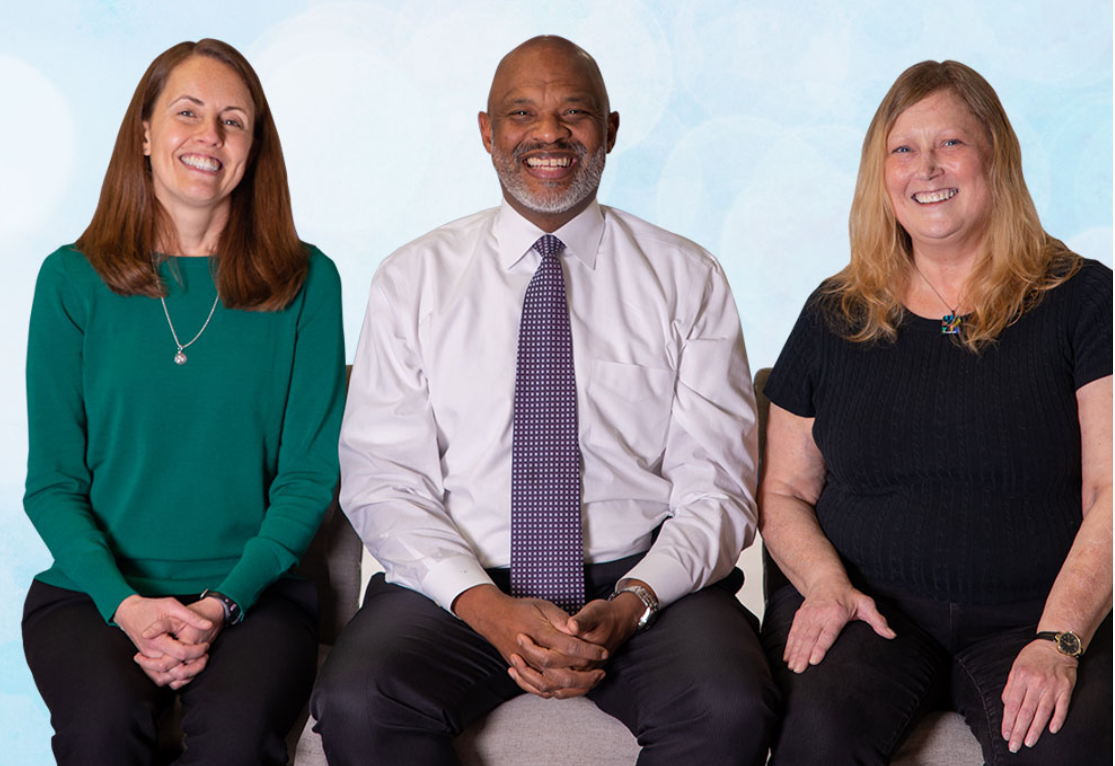 Three people seated: woman in green, man in white shirt, woman in black. Smiling, light blue background.