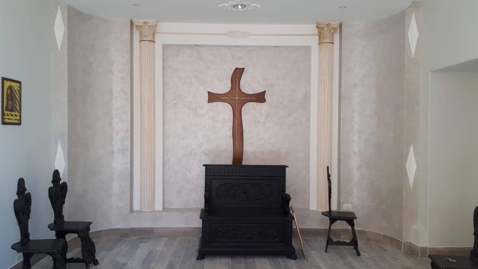 Interno di una cappella con una croce di legno su una cassa di legno scuro. Due sedie a sinistra e una a destra.