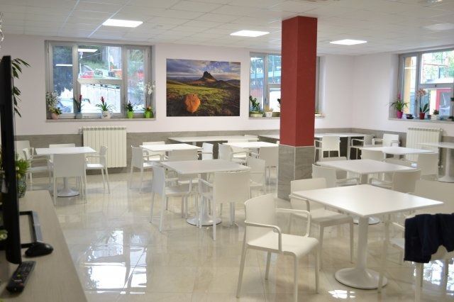 Caffetteria luminosa e moderna con tavoli e sedie bianchi e pavimento lucido. Ampie finestre, opere d'arte e una colonna di supporto rossa.
