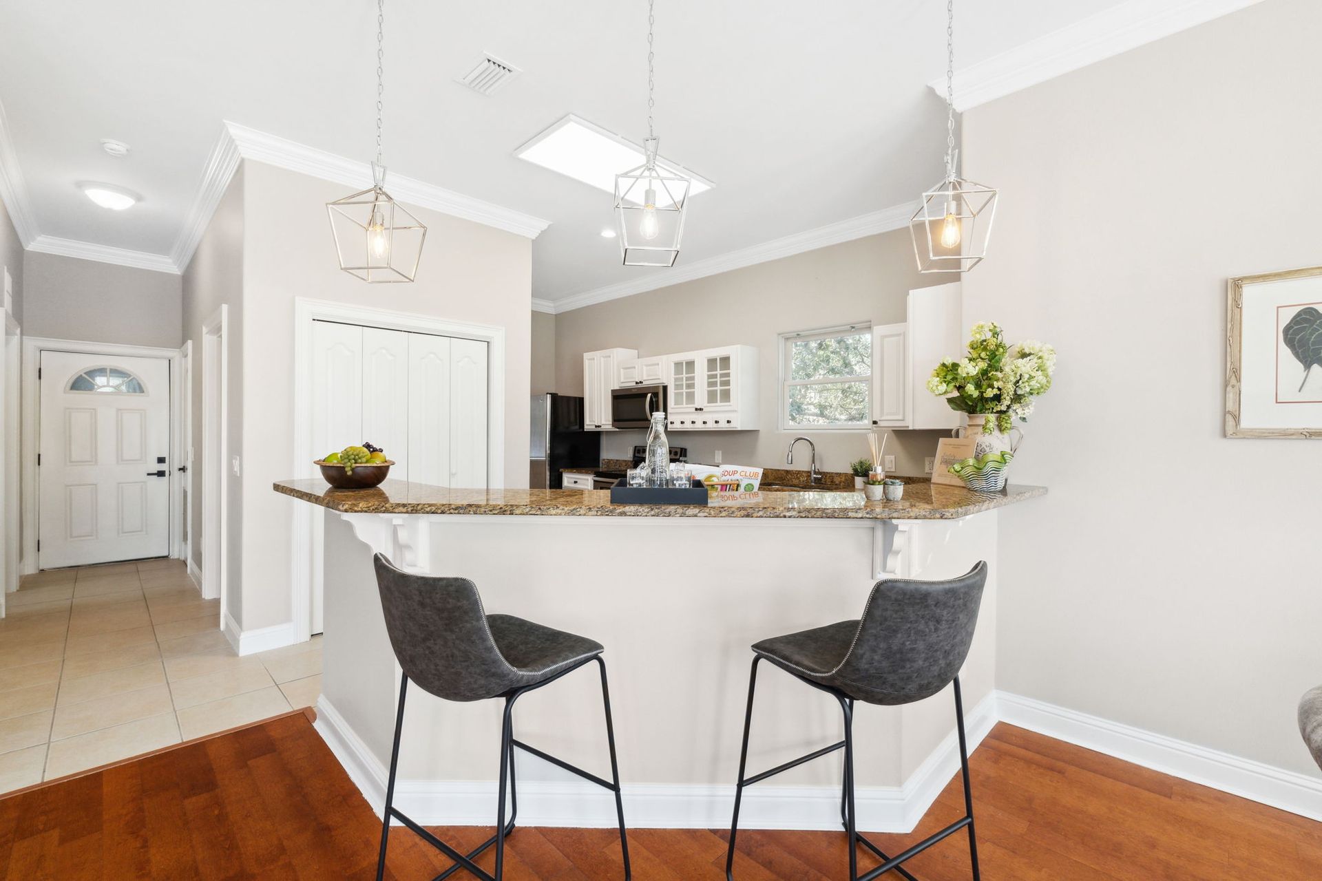 Bright modern kitchen with granite island, two black bar stools, and white cabinets in a hallway view