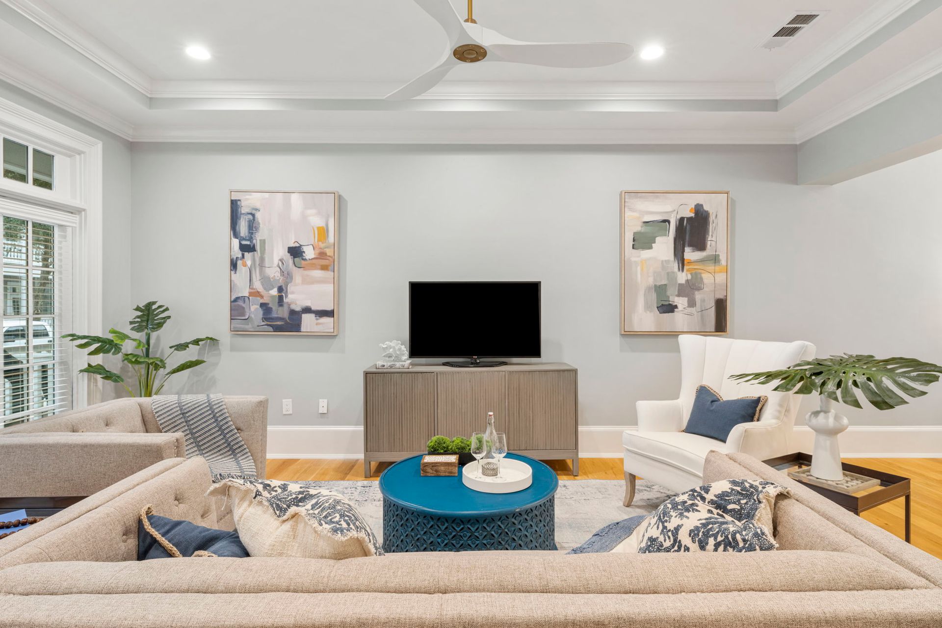 Bright modern living room with beige sofas, blue accents, TV, abstract art, and large windows