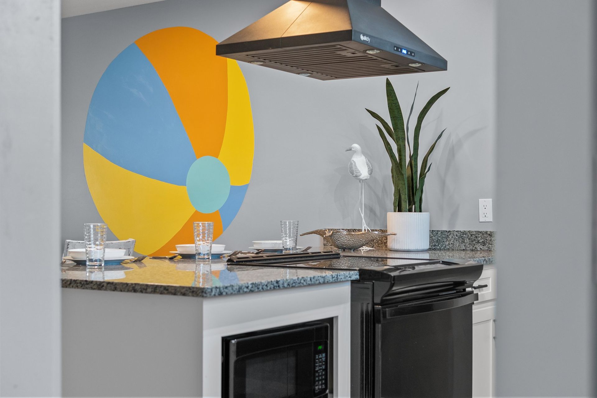 Modern kitchen with colorful wall art, granite countertop, stovetop, and a potted plant under a hood vent