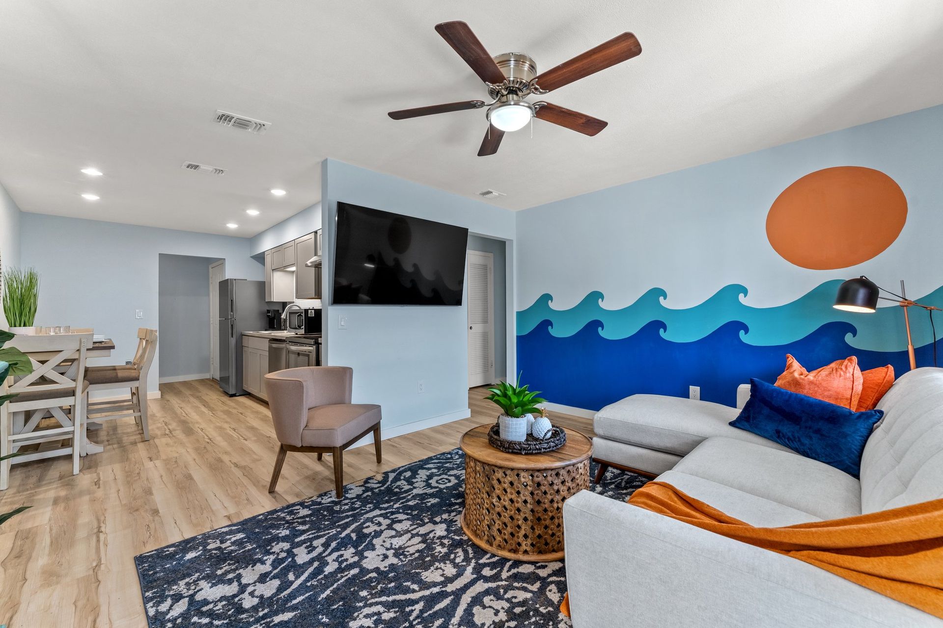 Bright coastal-style living room with blue wave mural, gray sofa, orange accents, and wall-mounted TV