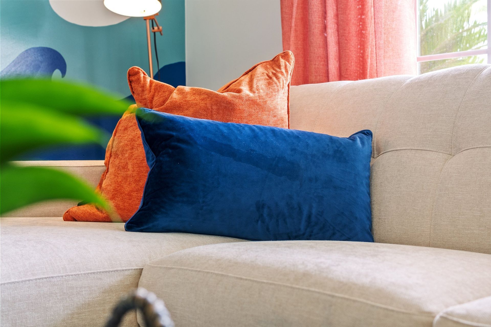 Beige sofa with orange and blue pillows beside a leafy plant and pink curtains