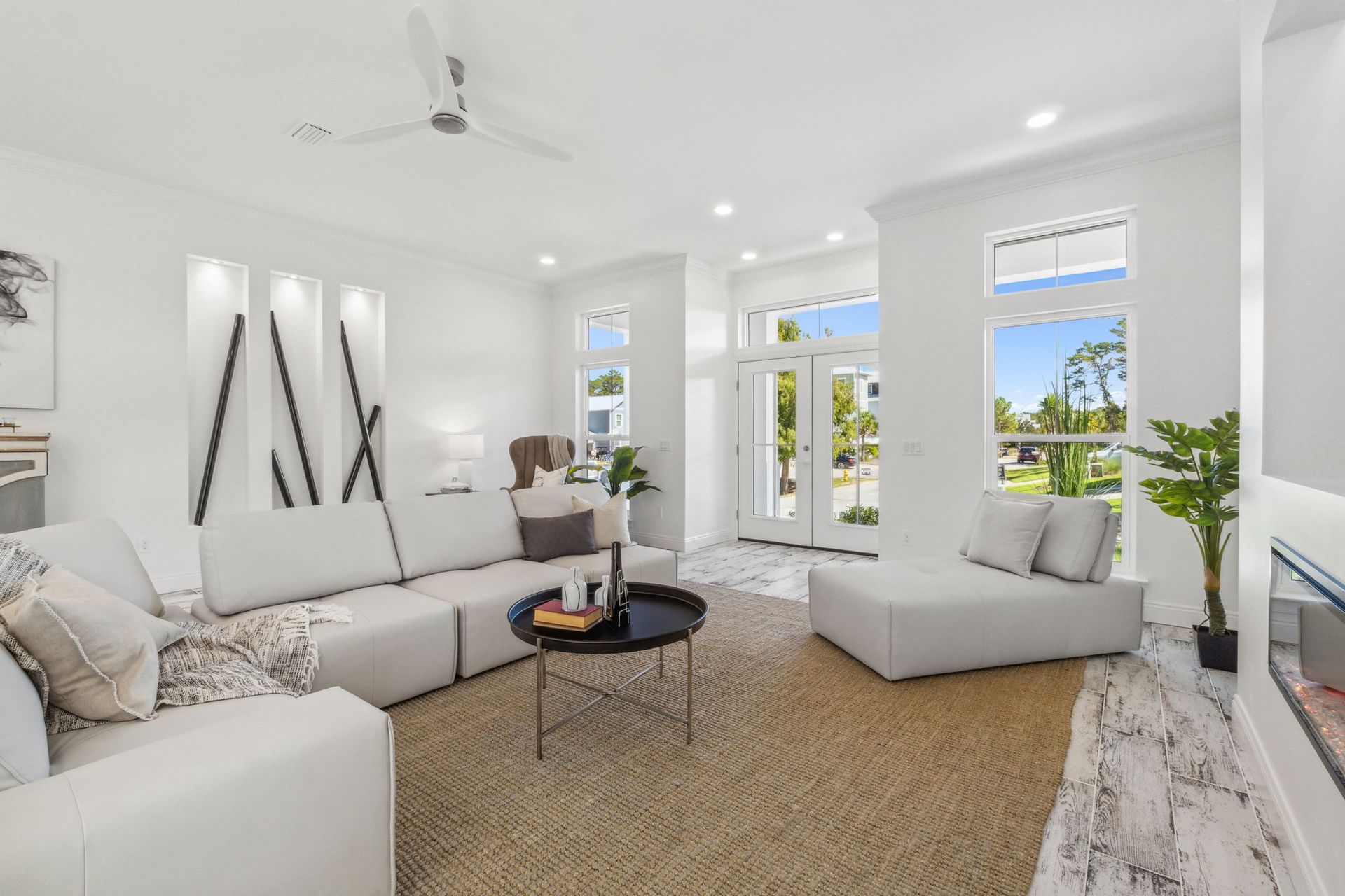 Bright white living room with sectional sofa, fireplace, woven rug, and large windows overlooking greenery