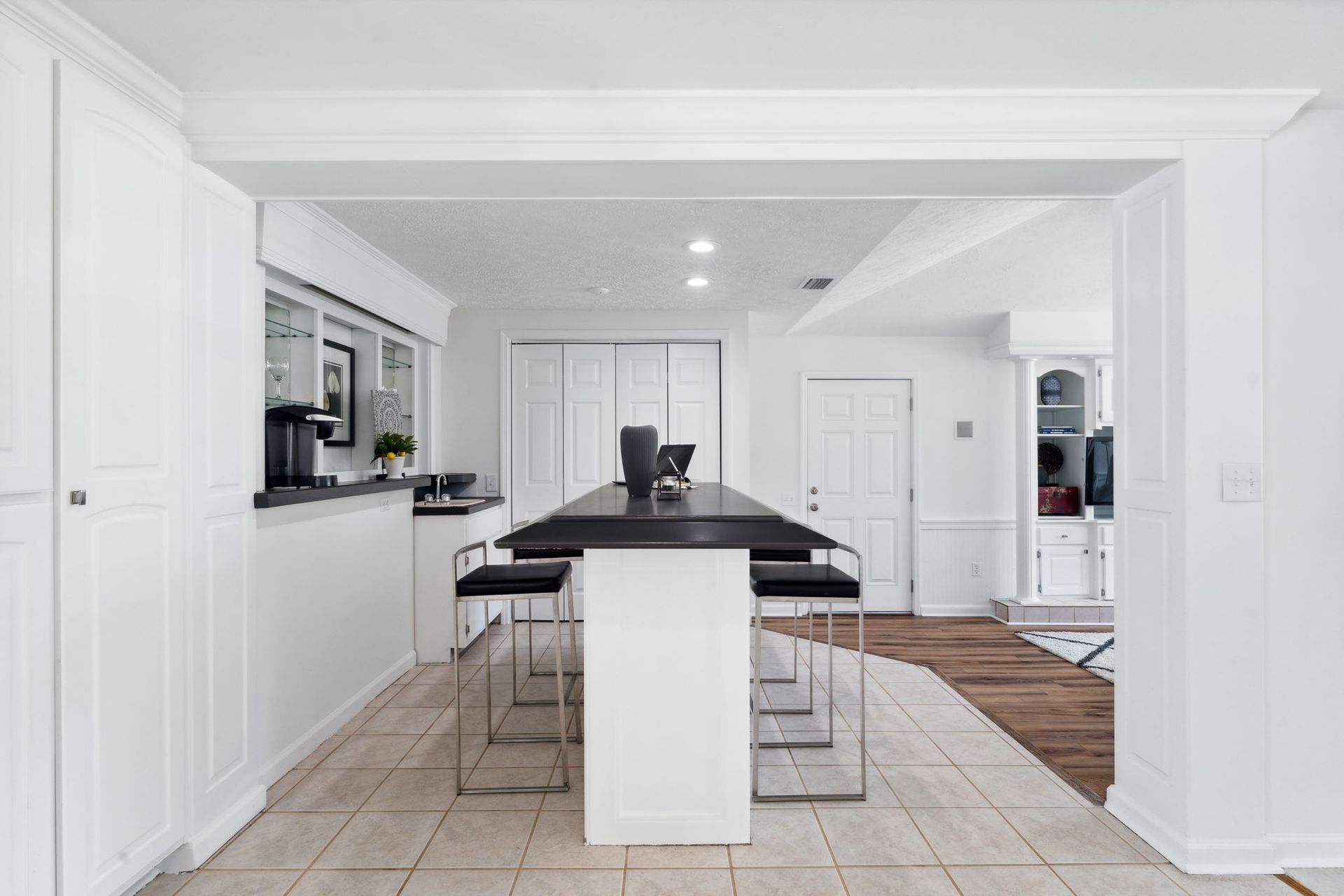 Bright white kitchen with a black island, bar stools, and tiled floor opening to a dining area