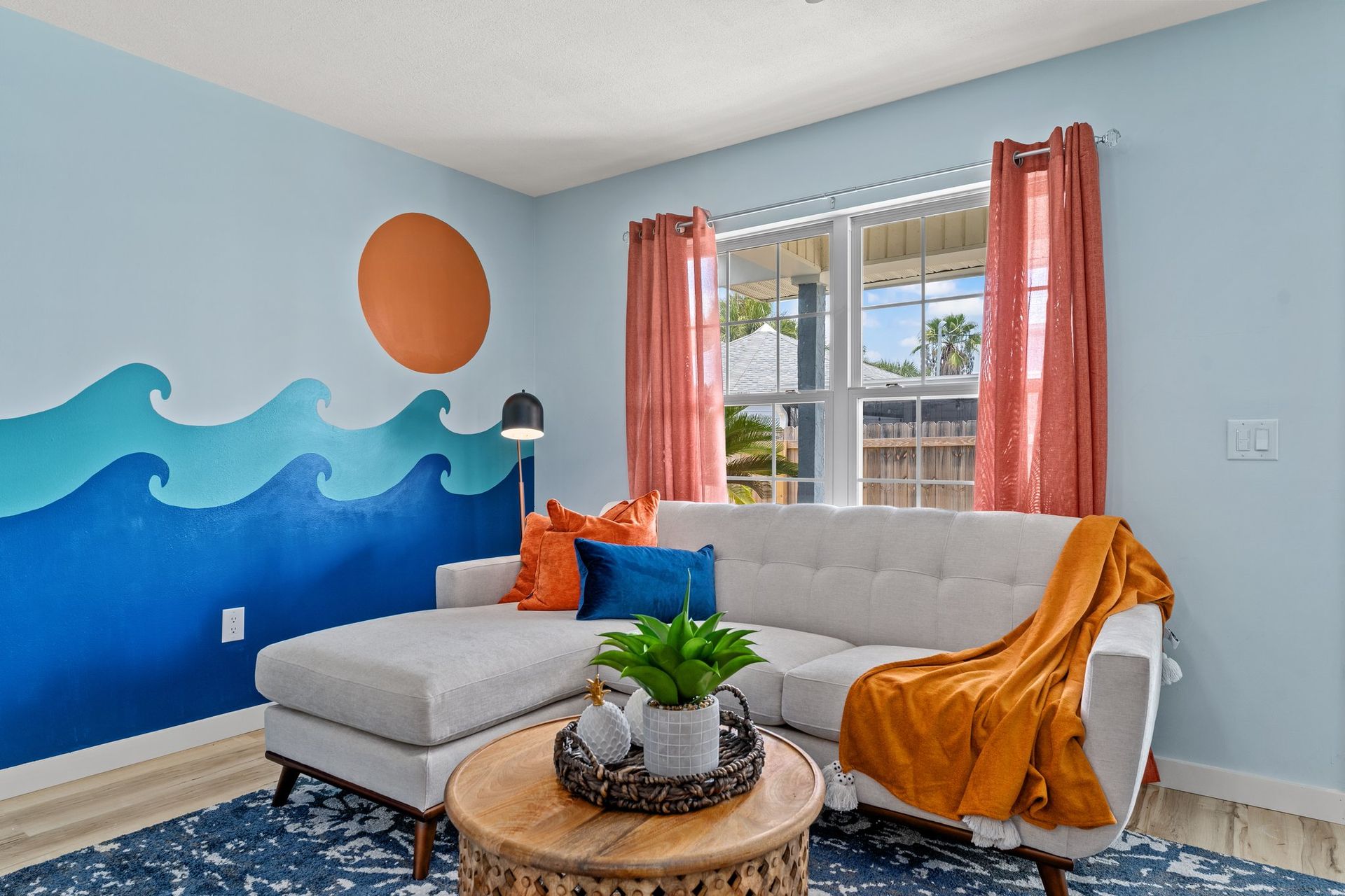 Bright living room with blue wave mural, white sectional, orange accents, and window with coral curtains