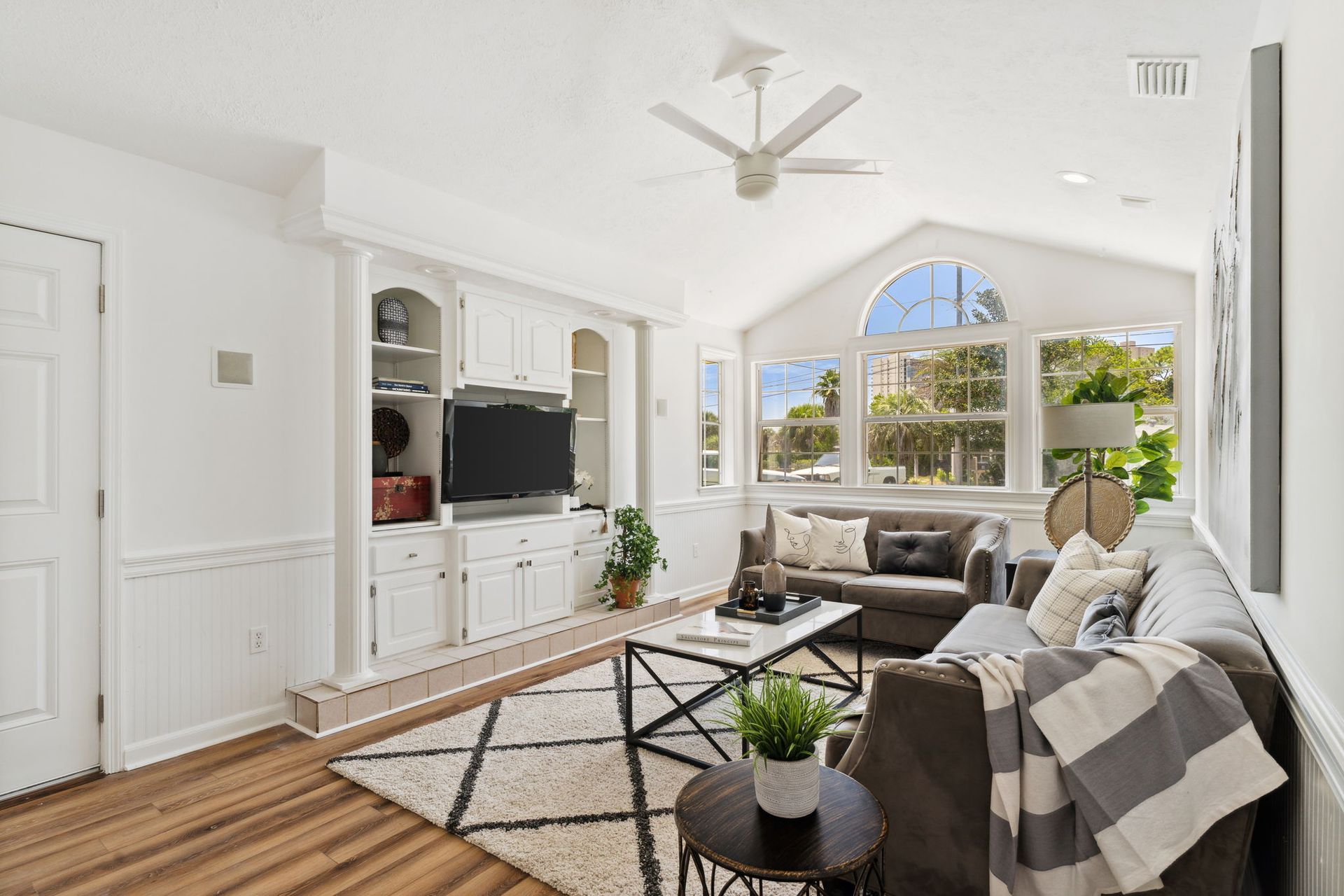 Bright living room with gray sofa, white built-ins, wood floors, and large arched windows overlooking greenery